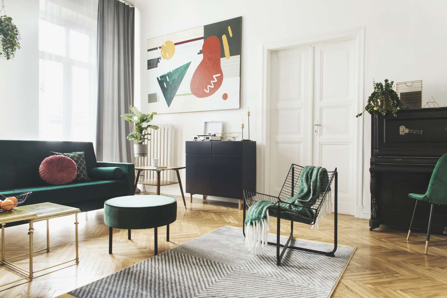 Solig lyxig heminredning med design sammetssoffa, fåtölj, bord, sittpuff, kommod och accessoarer. Fina växter på pianot. Vita väggar med målning. Stora fönster. Snygg inredning av vardagsrum
